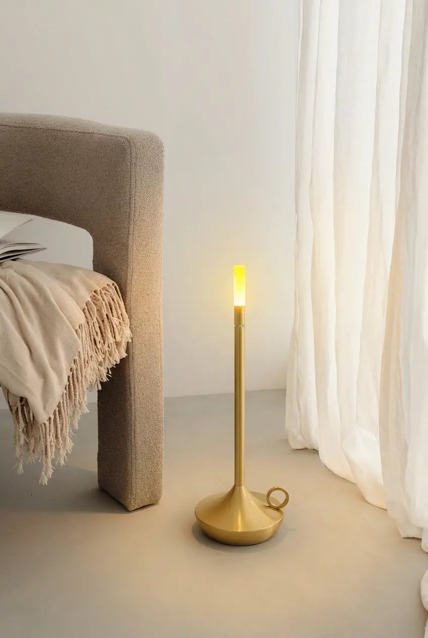 Wabi-sabi home corner: a matte gold witlity Aurora cordless table lamp with warm candle-like glowing light, its round base with a portable ring handle, rests on light gray microcement floor. Beside a beige fabric armchair with a fringed knit throw, soft natural light filters through sheer white linen curtains, creating a calm, cozy high-end home atmosphere.