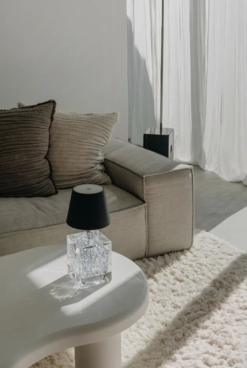 Minimalist cream living room corner: an ice-crack crystal table lamp with a witlity black bottle lamp rests on a white organic side table. A beige fabric sofa with textured knit pillows sits behind, while soft daylight through sheer curtains creates a calm, high-end modern home vibe with muted neutral tones.