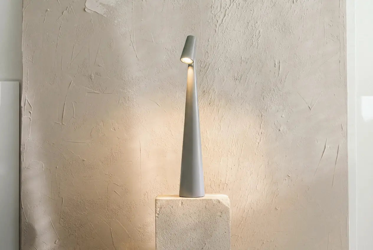 Gray witlity scalptower lamp, with an angled lamp head casting warm soft downward light, its sleek streamlined body rests on a beige travertine-textured square pedestal. Against a textured gray-beige art paint wall, the warm glow creates soft shadows, bringing a calm, high-end wabi-sabi minimalist home atmosphere.