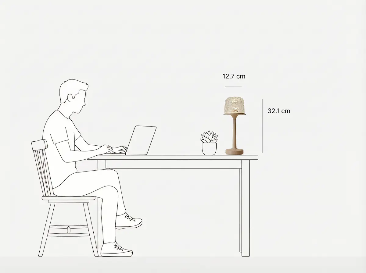 Minimalist black-and-white line art showing a table lamp's usage scene and size specs. A man sits on a wooden chair working on a laptop at a desk, with a small succulent and the Witlity's skywave lamp on the table. Marked with 32.1 cm total height and 12.7 cm shade diameter, it clearly presents the lamp's size proportion and desktop compatibility.