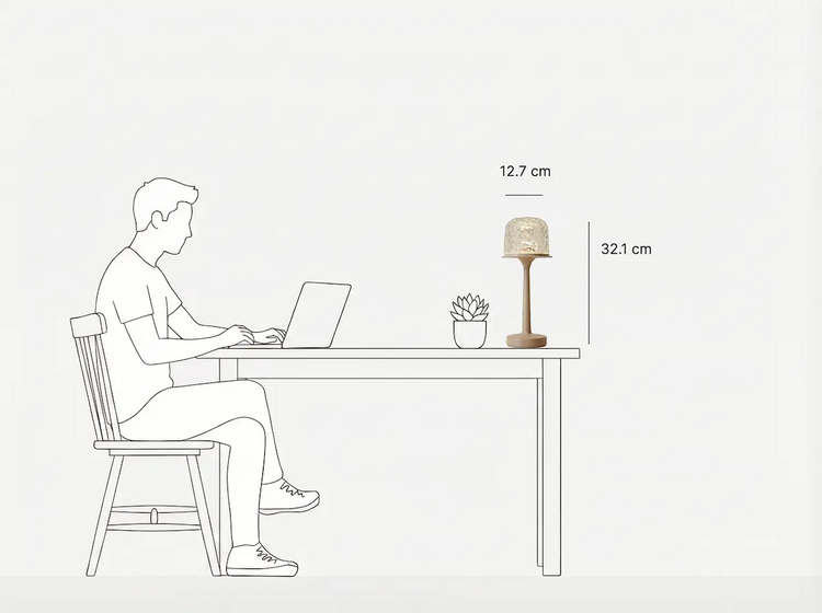 Minimalist black-and-white line art showing a table lamp's usage scene and size specs. A man sits on a wooden chair working on a laptop at a desk, with a small succulent and the Witlity's skywave lamp on the table. Marked with 32.1 cm total height and 12.7 cm shade diameter, it clearly presents the lamp's size proportion and desktop compatibility.
