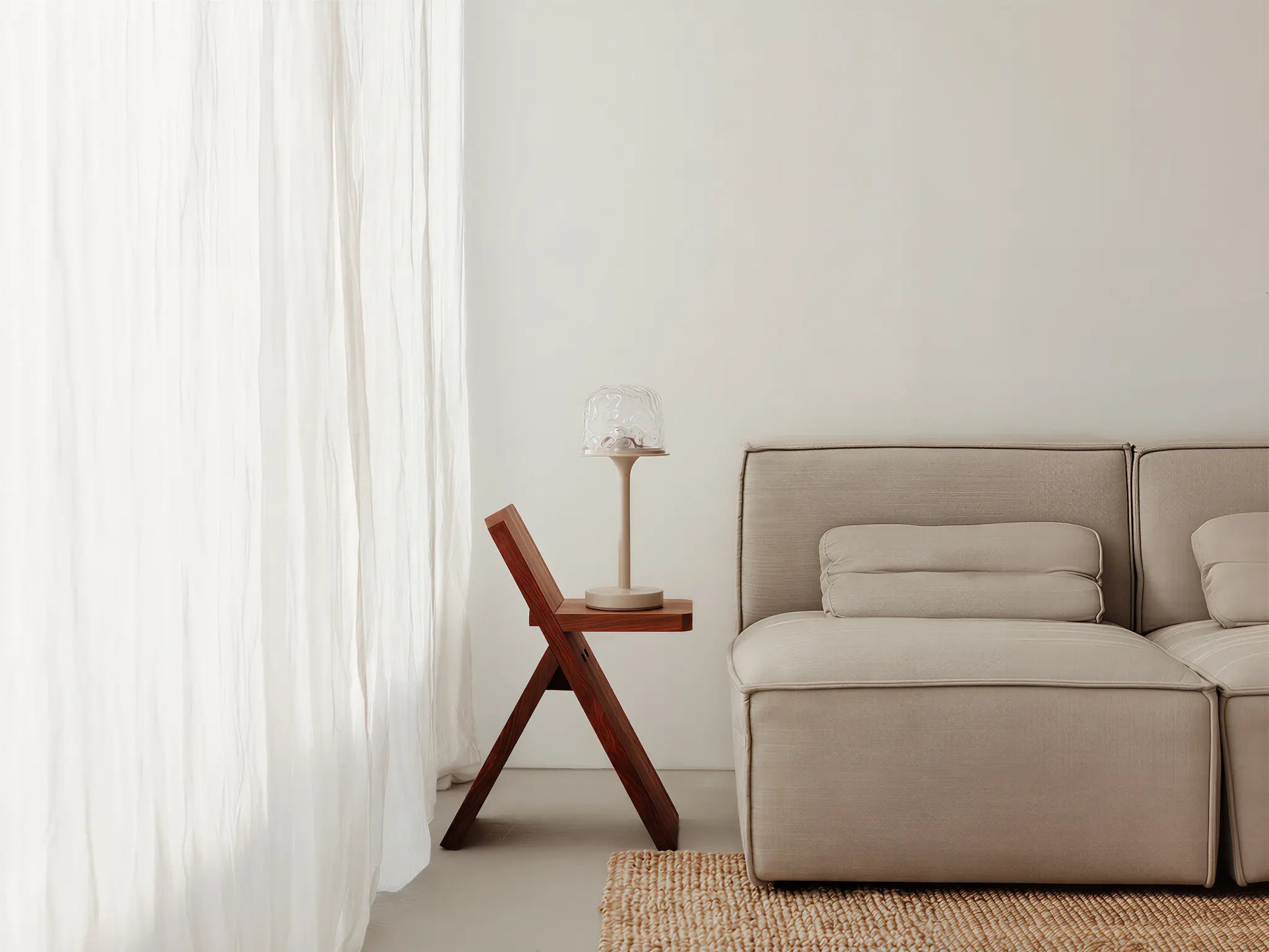 Minimalist cream-style home scene: a witlity skywave table lamp with rippled transparent glass shade and matte beige body rests on a dark walnut folding solid wood side table. Beside a light beige fabric modular sofa with matching cushions, soft daylight filters through sheer white crinkled curtains over a natural jute rug, creating a warm, clean high-end home atmosphere.